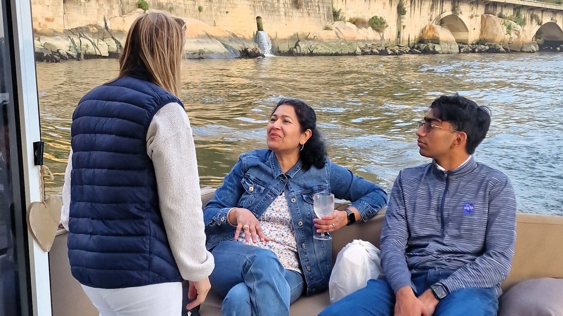 Three people talking on a boat near a stone bridge over a river.