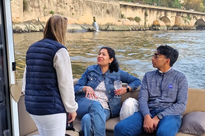 Three people talking on a boat near a stone bridge over a river.