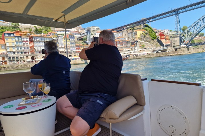 Two people on a boat take photos of a colorful riverside city with a bridge.