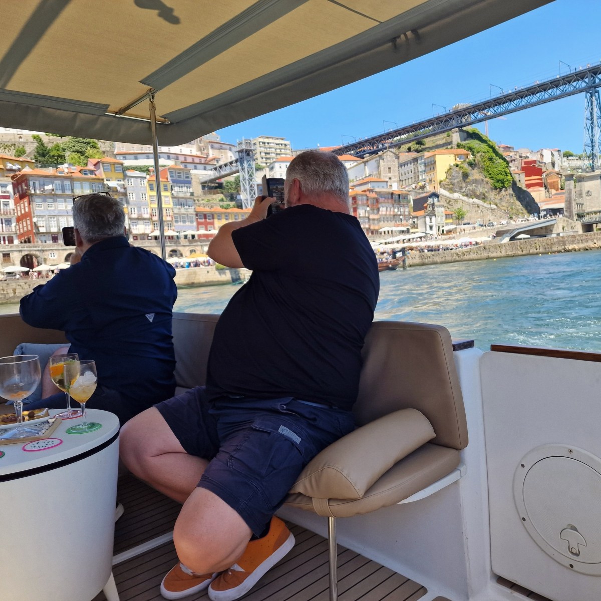 Two people on a boat take photos of a colorful riverside city with a bridge.