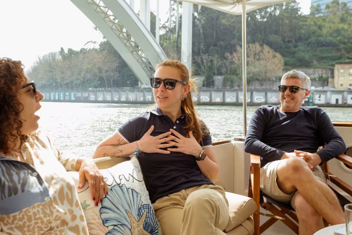 Three people wearing sunglasses chatting and laughing on a boat under a bridge.