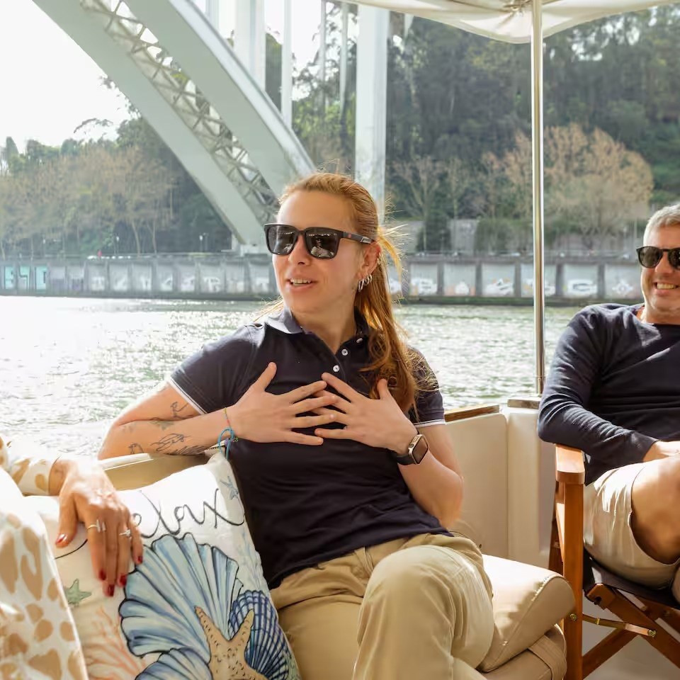 Three people wearing sunglasses chatting and laughing on a boat under a bridge.