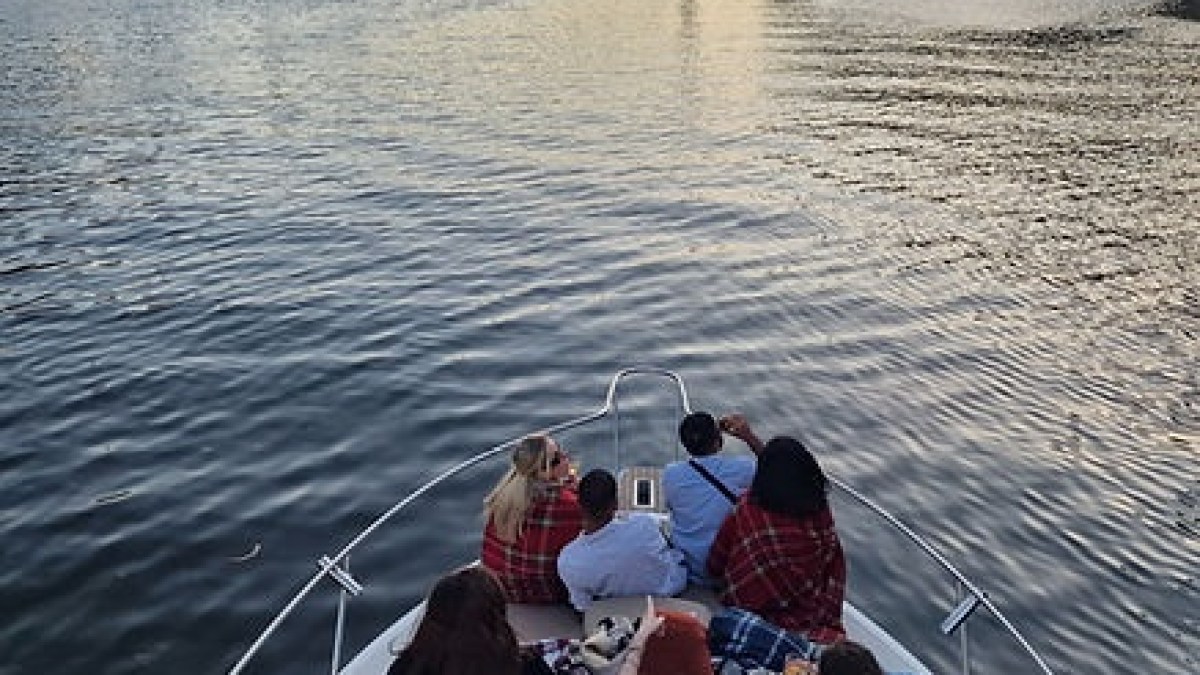 Group of people on a boat in a river heading towards a bridge at sunset.
