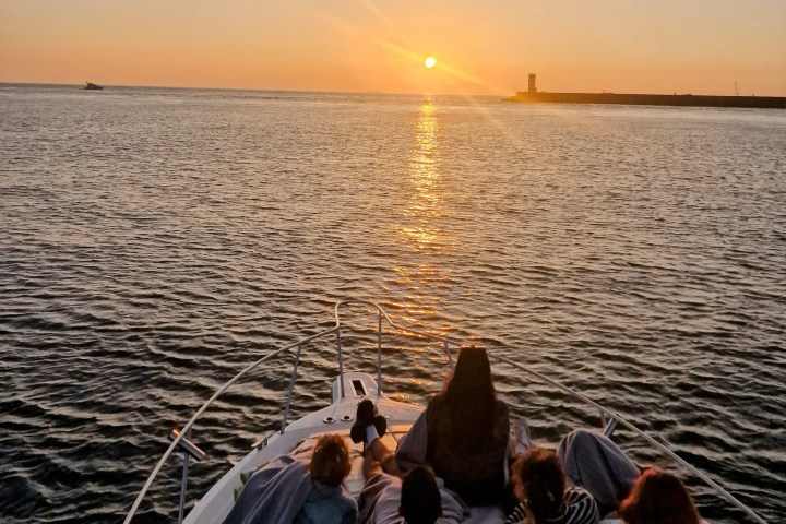 People on a boat watching a sunset over the ocean with a distant shoreline.