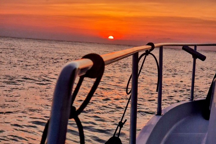 Boat railing with sunset over ocean and cloudy sky in background.