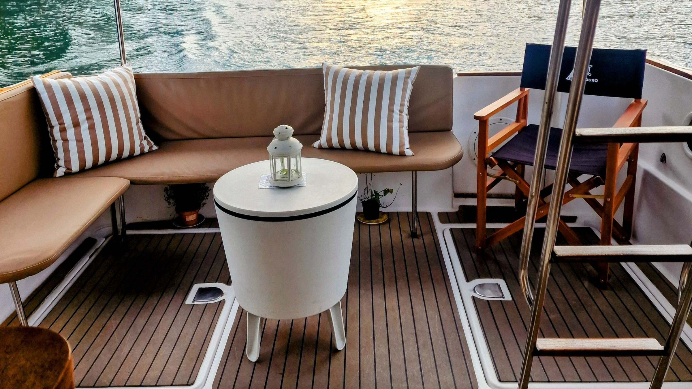 Boat deck with seating, striped cushions, table, lantern, and view of water at sunset.