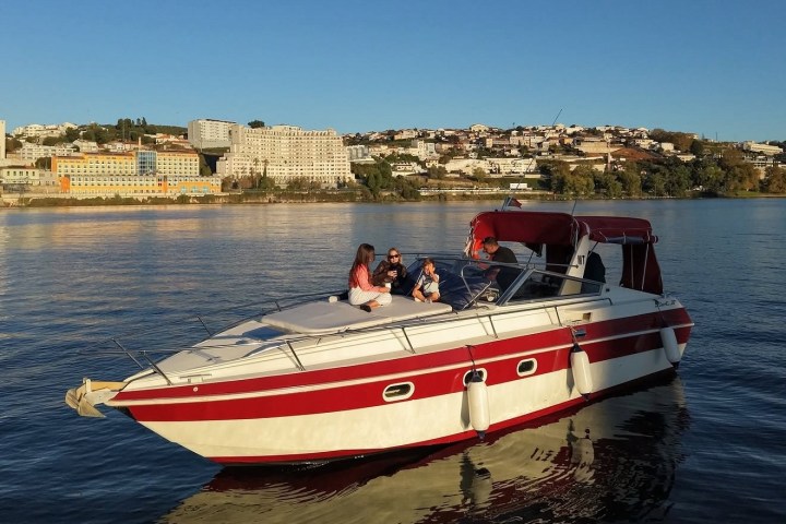 People sitting on a red and white boat in calm water, with a cityscape in the background.