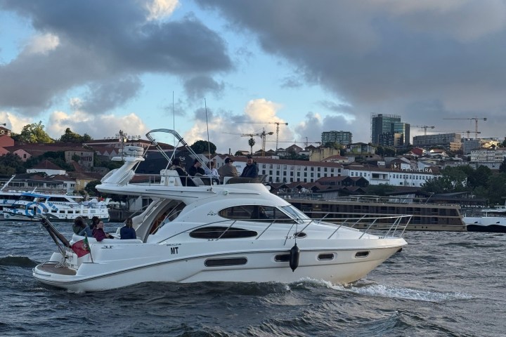White yacht with people on a wavy river, cityscape in background under cloudy sky.