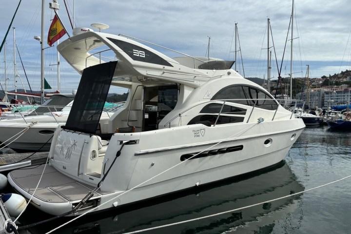 White yacht docked in a marina with other boats under a cloudy sky.